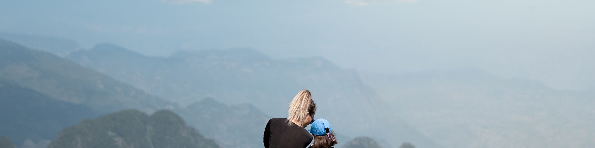 Uma mulher e uma criança sentadas numa colina gramada, com vista para uma cordilheira.