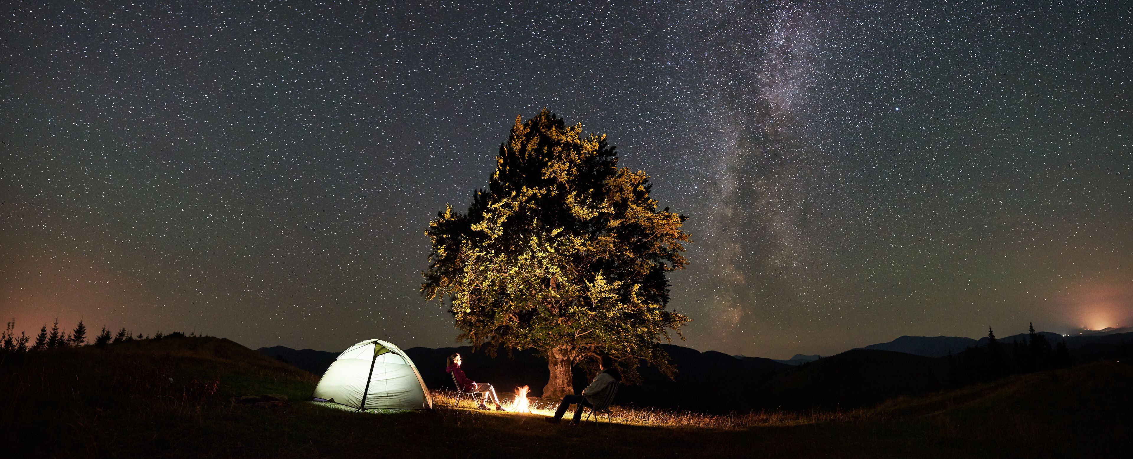 Panoramic view of couple tourists resting at summer night camping in mountains. Travelers sitting on chairs beside campfire, illuminated tent and big tree under starry sky full of stars and Milky way.