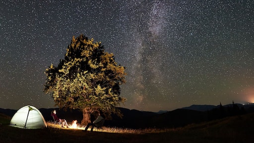 Panoramic view of couple tourists resting at summer night camping in mountains. Travelers sitting on chairs beside campfire, illuminated tent and big tree under starry sky full of stars and Milky way.