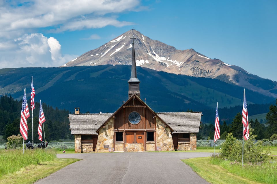 Een stenen kapel met een kruis, omringd door Amerikaanse vlaggen, voor een berg.