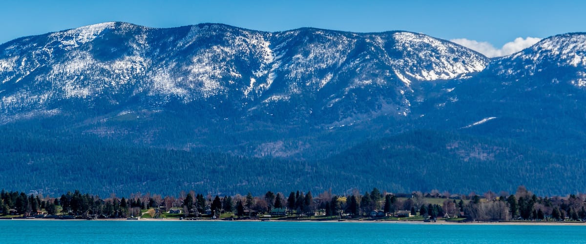A mountain range with snow-capped peaks, a clear blue sky, and a shoreline with trees and buildings.