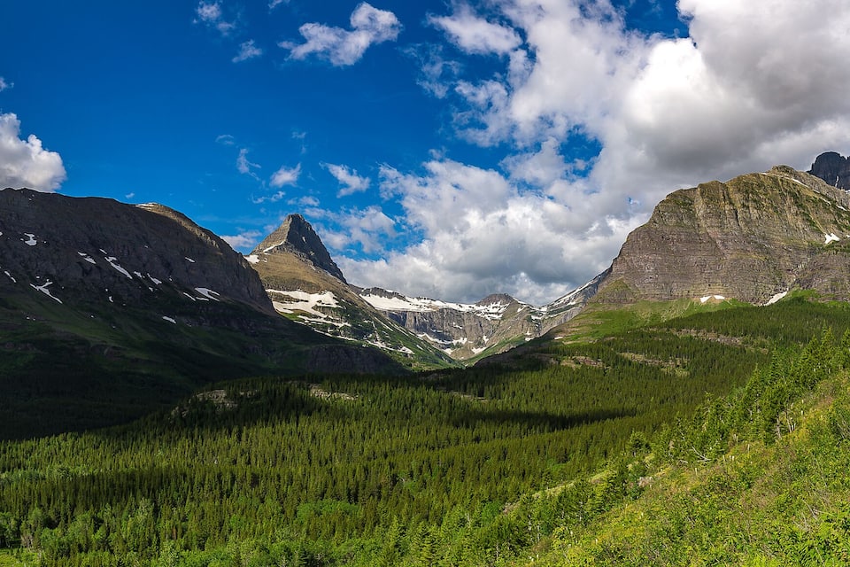 A mountain landscape with a dirt path, dense forest, and snow-capped peaks under a partly cloudy sky.