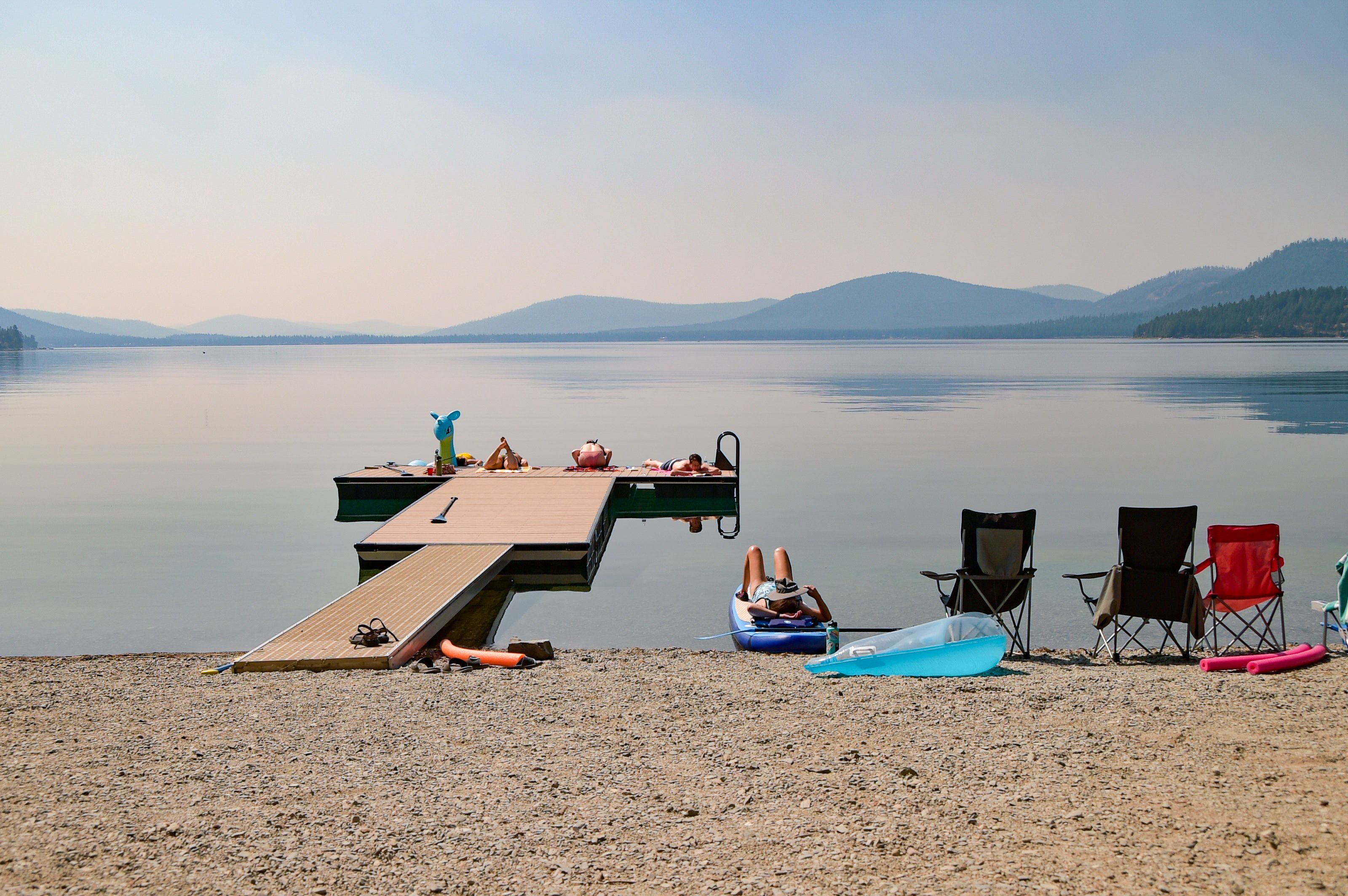A calm lake with a wooden dock extending into the water, surrounded by mountains and clear skies.