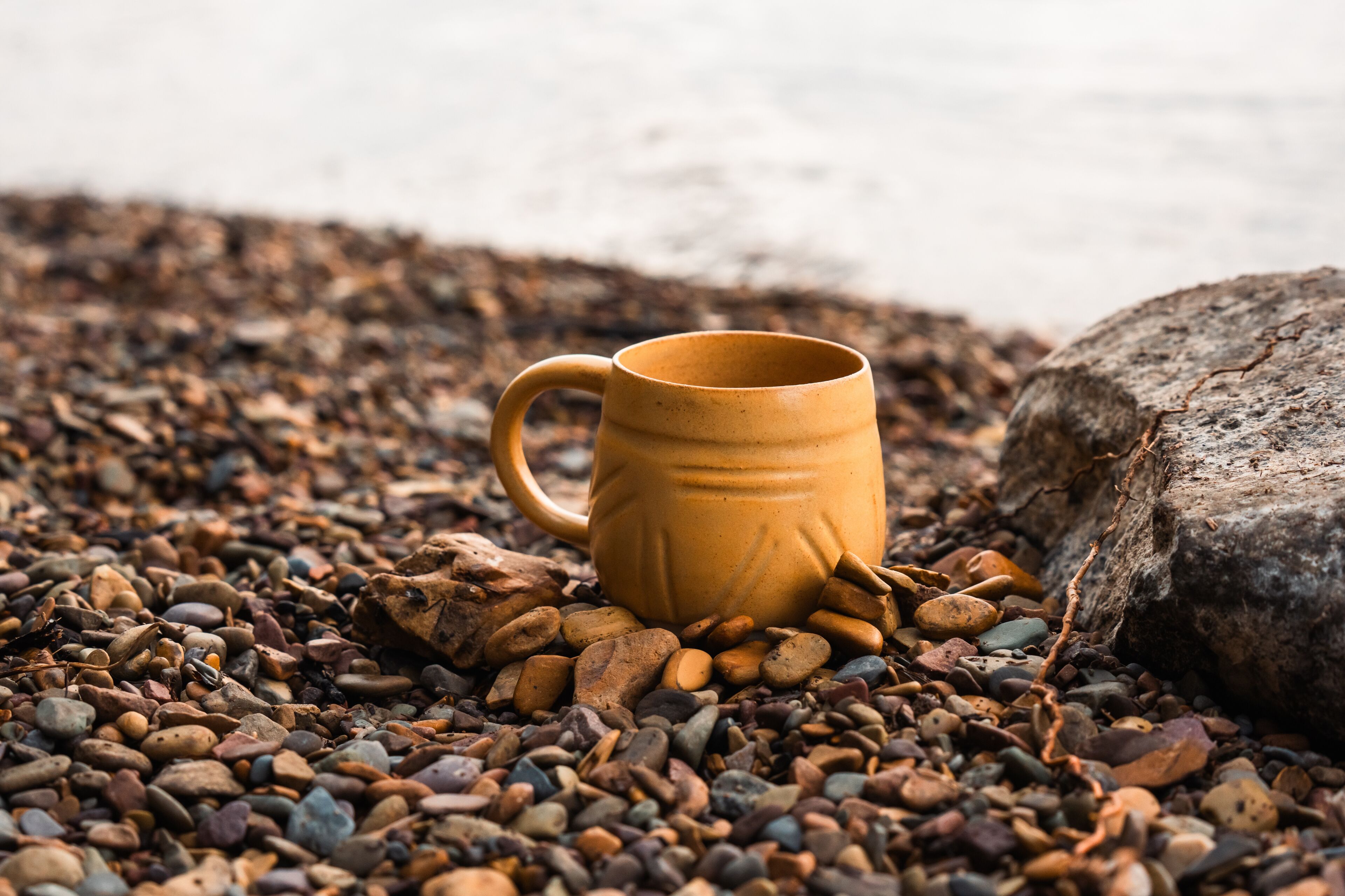 A yellow ceramic mug on a bed of multicolored pebbles with a large rock nearby.