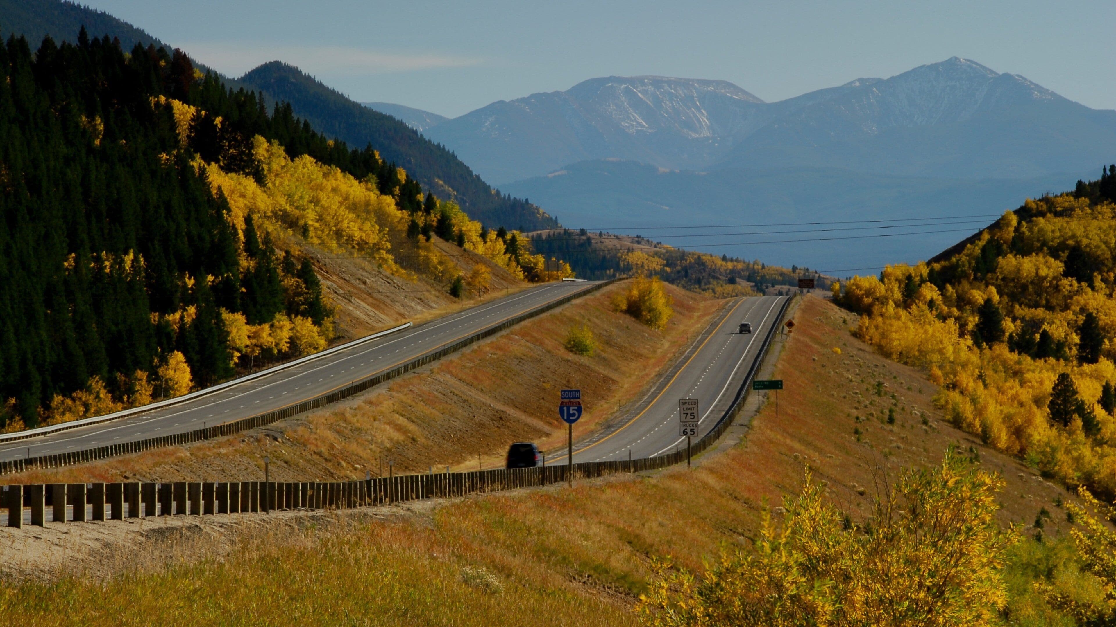 A highway curving through a mountainous landscape with autumn foliage and distant snow-capped mountains.