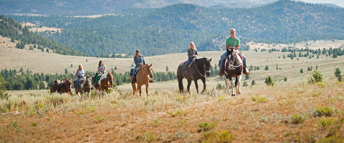 A group of people horseback riding across a grassy field with mountains in the background.
