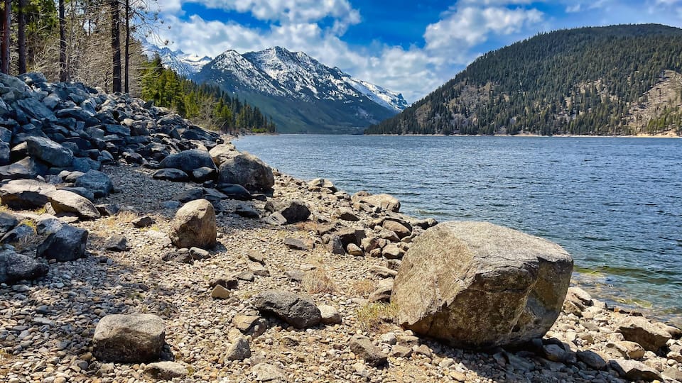 A rocky shoreline with a lake, mountains, and a forested area.