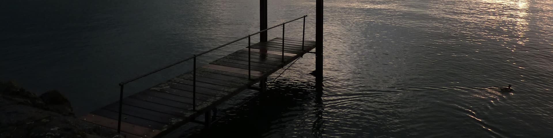 A calm lake with a wooden dock, mountains in the background, and the sun setting on the horizon.
