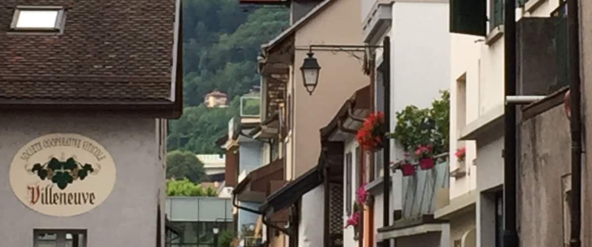 A narrow street with buildings, a mountain in the background, and a sign that reads "Société Coopérative Viticole Villeneuve".