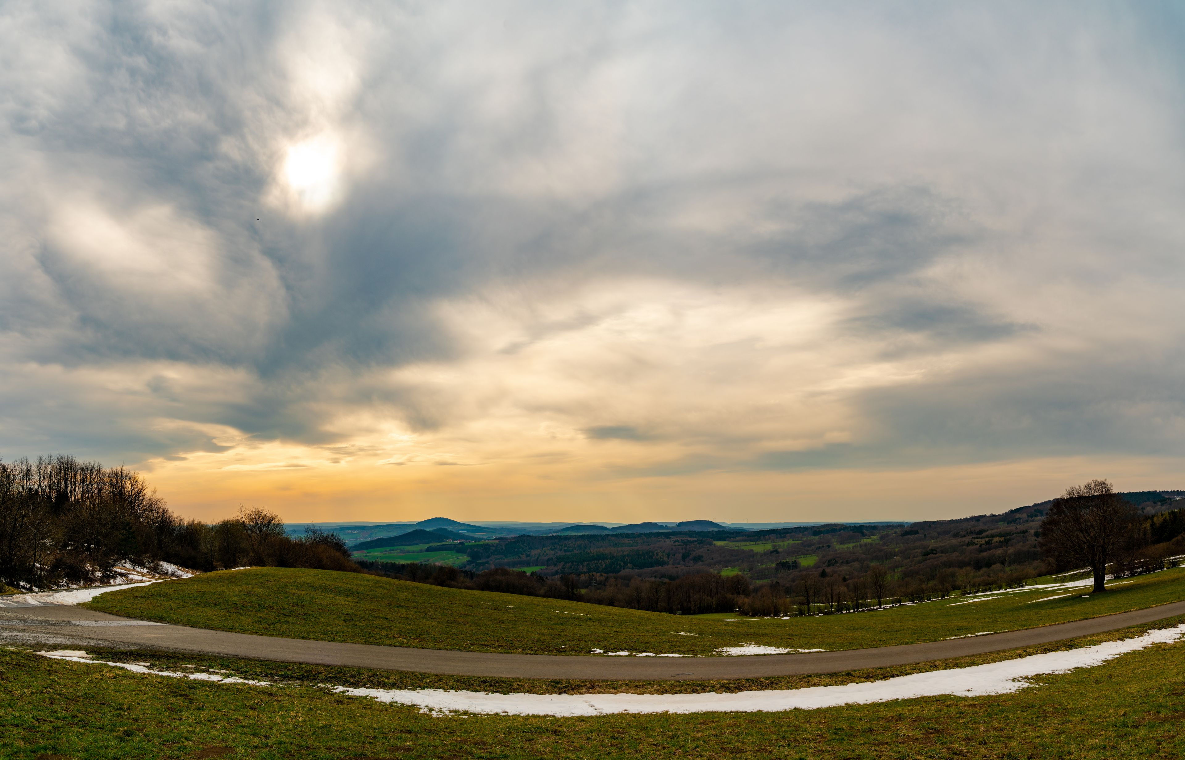 A landscape with a winding road, snow patches, and a cloudy sky.