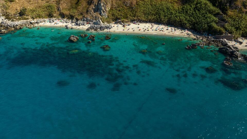 An aerial view of a turquoise beach with clear waters, rocky outcrops, and a forested hillside.