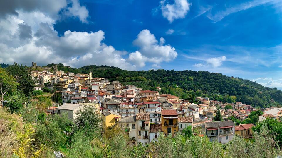 A hillside village with terracotta-roofed buildings, lush greenery, and a prominent castle-like structure in the background.