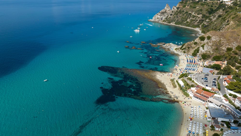 Eine Küstenstadt mit klarem türkisblauem Wasser, felsigen Klippen und einem Strand mit Sonnenschirmen und Booten.