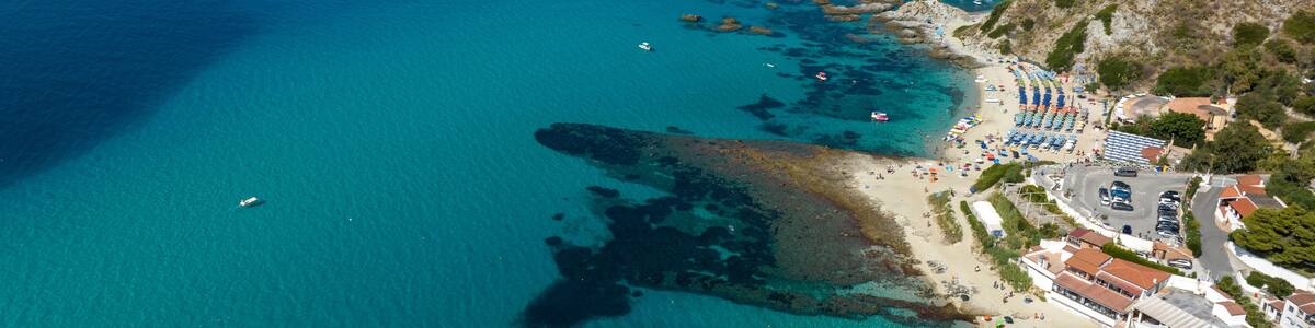 A coastal town with clear turquoise waters, rocky cliffs, and a beach with umbrellas and boats.