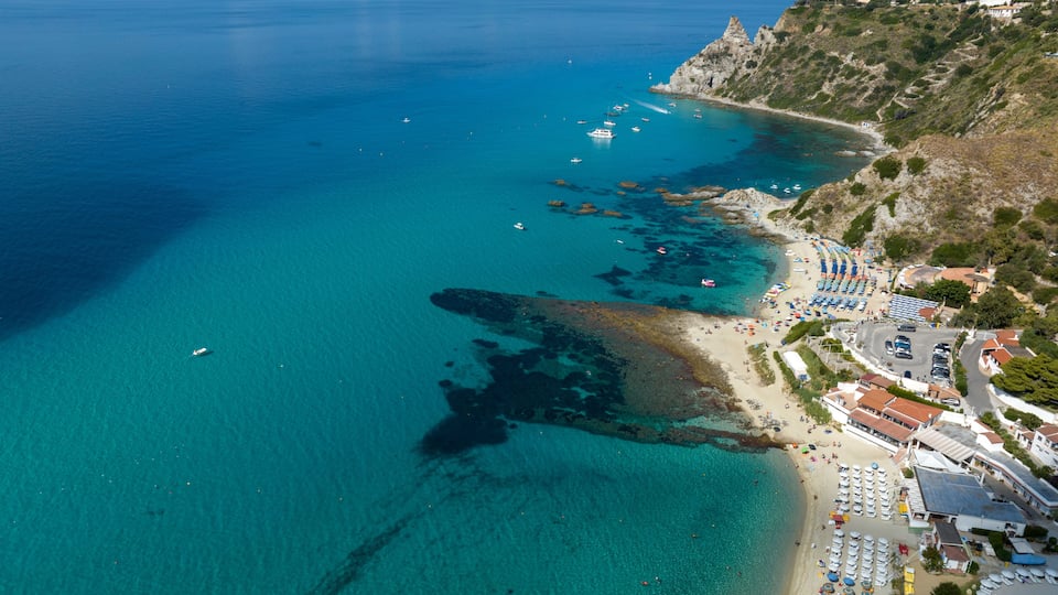 A coastal town with clear turquoise waters, rocky cliffs, and a beach with umbrellas and boats.