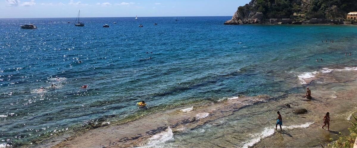 A coastal scene with clear blue water, boats, and people swimming and walking on the shore.