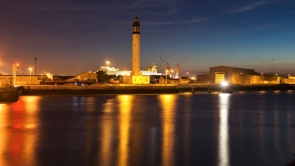 A lighthouse at dusk with a calm river reflecting lights, a construction crane, and a building with illuminated windows.