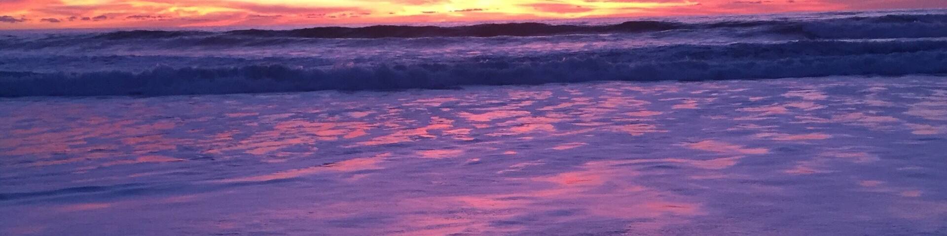 A beach at sunset with waves and a pink sky.