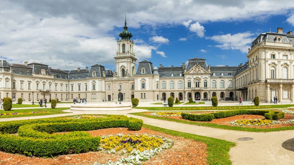 A grand historic building with a central tower, surrounded by well-manicured gardens and pathways.