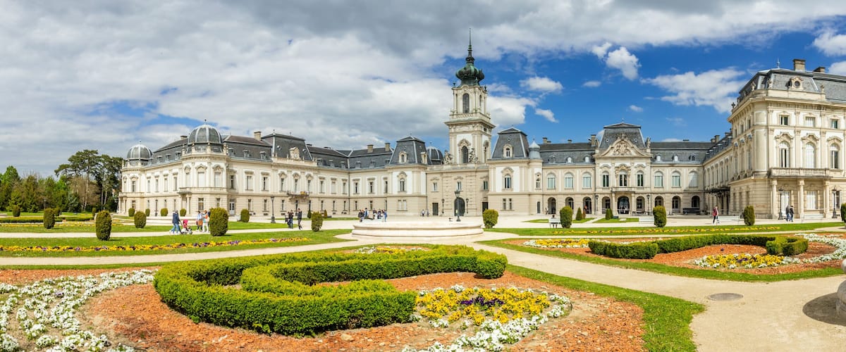A grand historic building with a central tower, surrounded by well-manicured gardens and pathways.