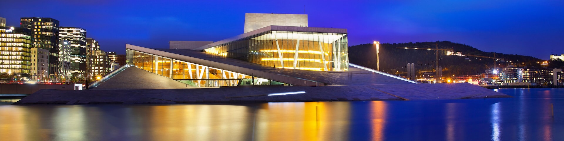 Um edifício moderno com arquitetura marcante, janelas de vidro refletivas, uma orla marítima e paisagem urbana ao entardecer.