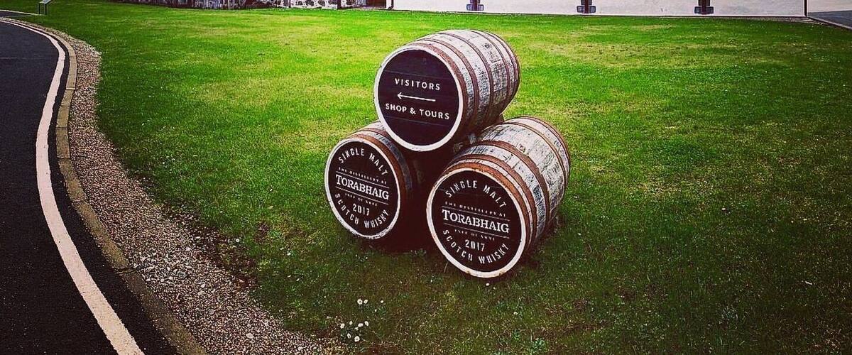 Un edificio con la leyenda 'TORABHAIG' que presenta un muro de piedra y una zona con césped. Tres barriles con la inscripción 'TORABHAIG SINGLE MALT SCOTCH WHISKEY' están en primer plano.