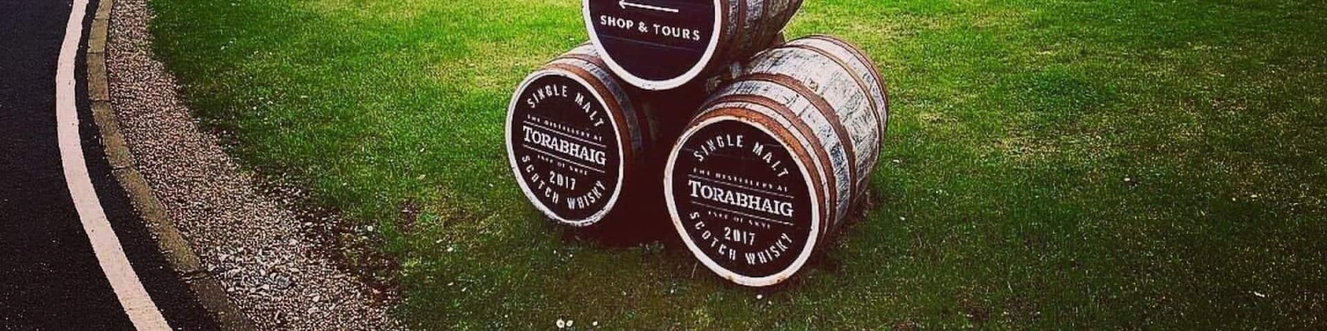 A building labeled 'TORABHAIG' with a stone wall and a grassy area. Three barrels with 'TORABHAIG SINGLE MALT SCOTCH WHISKEY' are in the foreground.