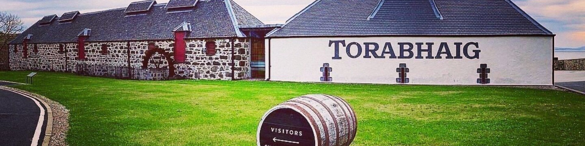 A building labeled 'TORABHAIG' with a stone wall and a grassy area. Three barrels with 'TORABHAIG SINGLE MALT SCOTCH WHISKEY' are in the foreground.