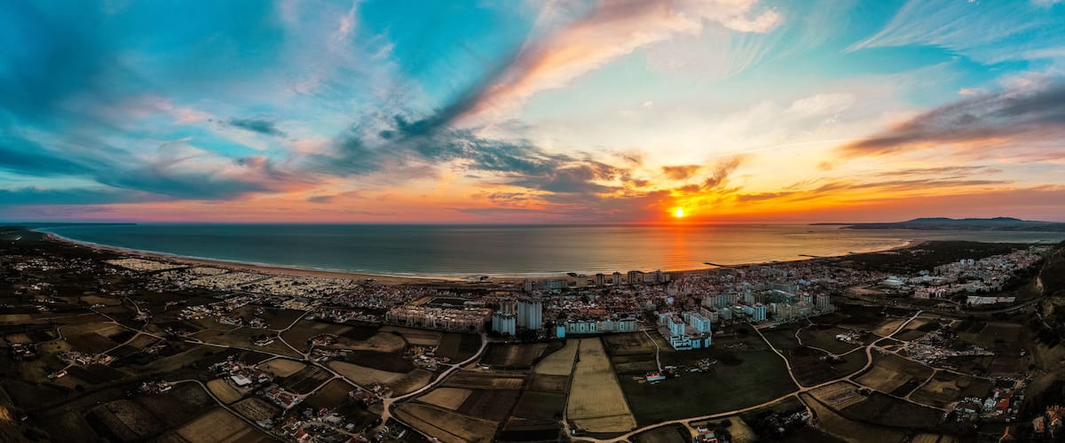 A coastal cityscape at sunset with a vibrant sky, a beach, and a mix of residential and commercial buildings.
