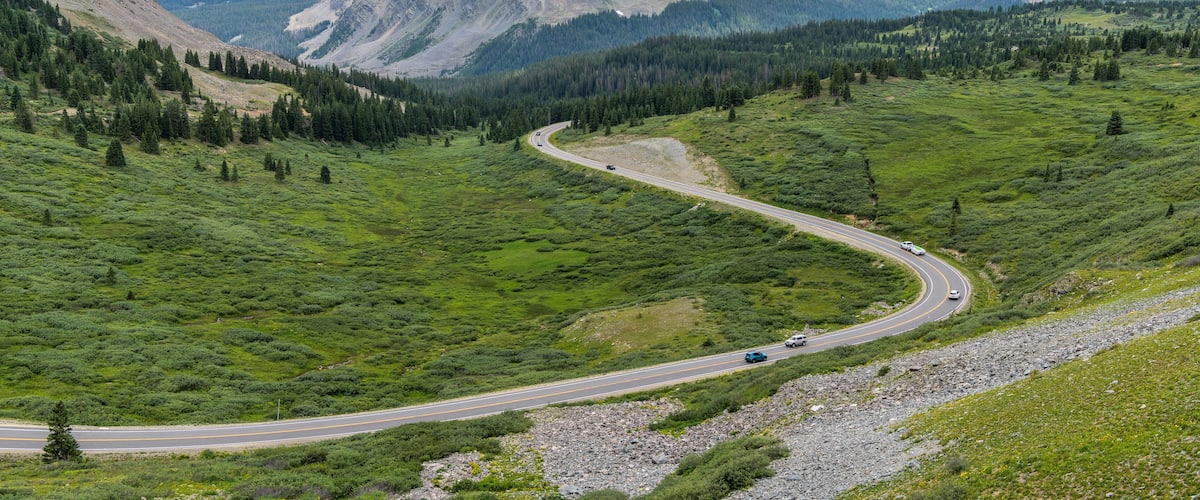 A winding mountain road through a lush green valley with forested hills in the background.