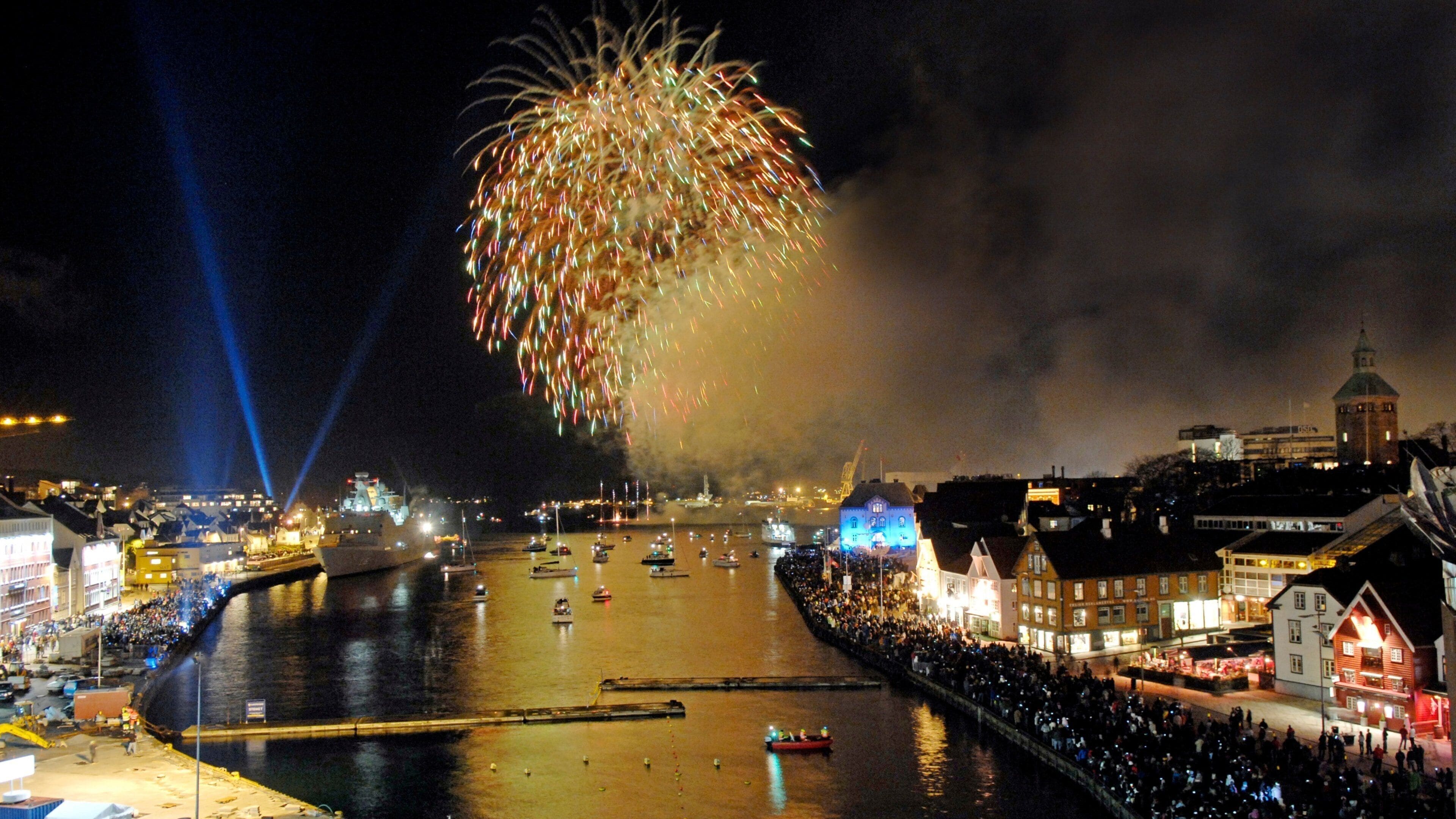 Una ciudad junto al agua con fuegos artificiales sobre el río, barcos y una multitud de gente.