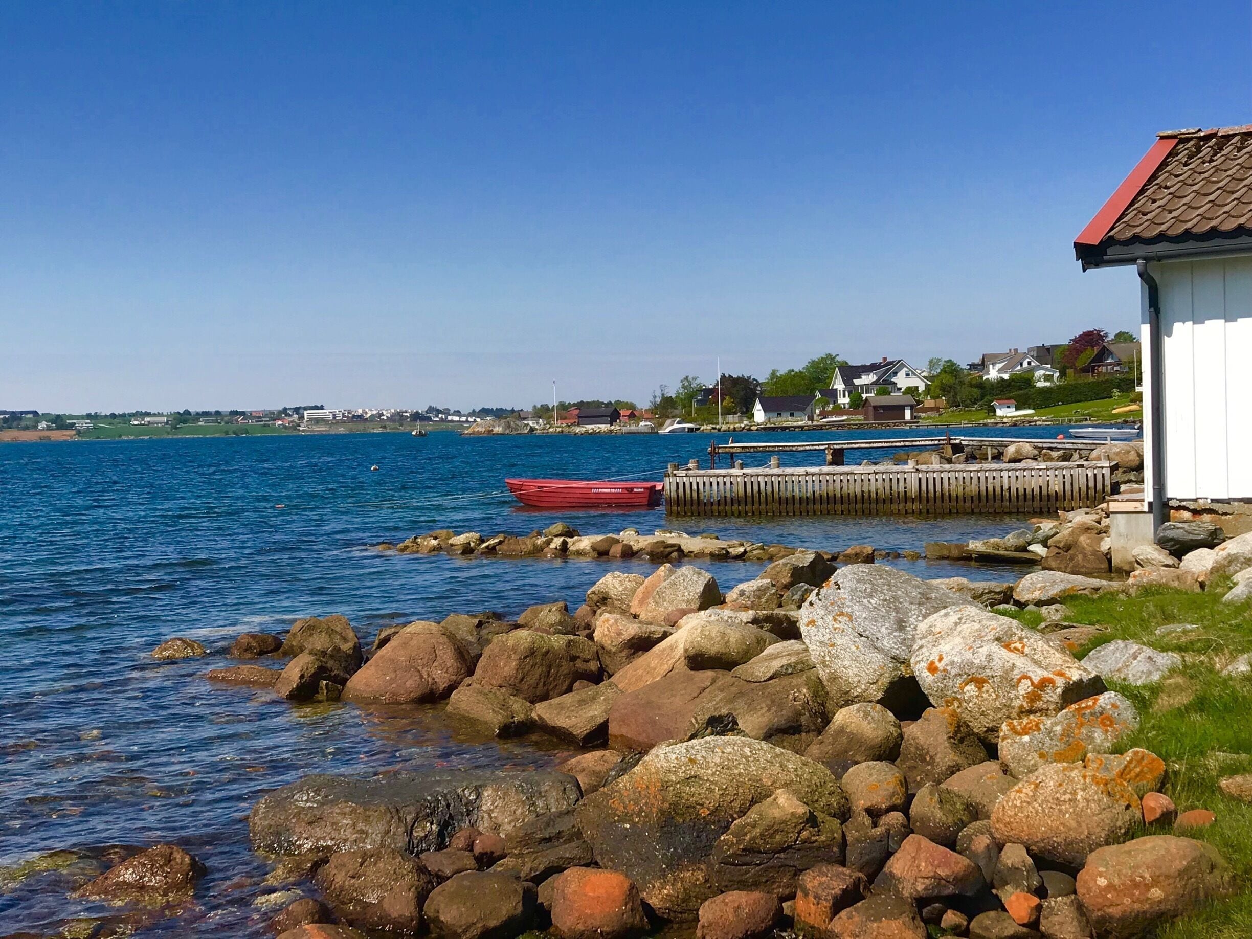 A rocky shoreline with a red boat docked at a wooden pier, a white building with a red roof, and houses in the background.