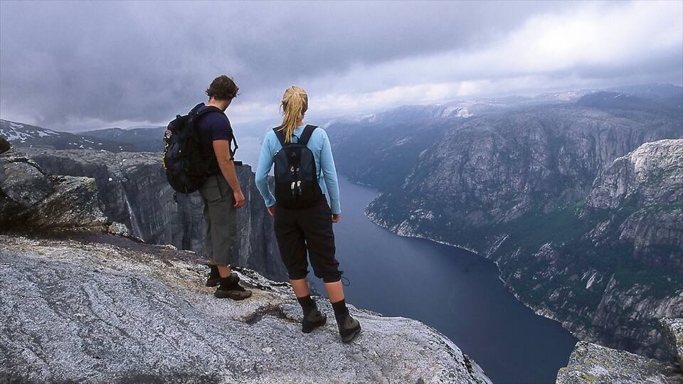 Zwei Wanderer auf einer felsigen Klippe mit Blick auf ein tiefes Tal, einen See und Berge.