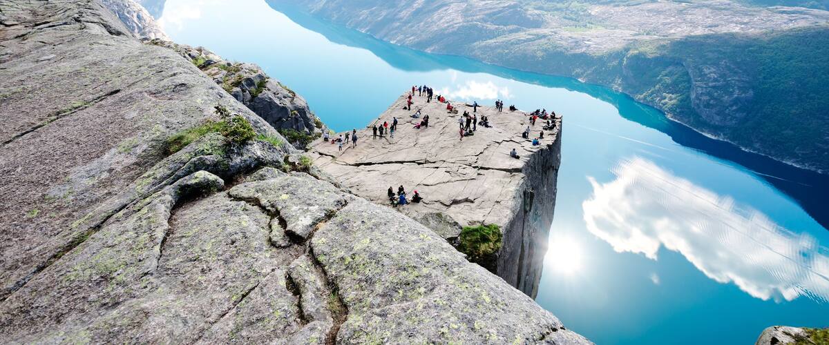 A rocky cliff with a group of people at the edge overlooking a turquoise lake surrounded by mountains.