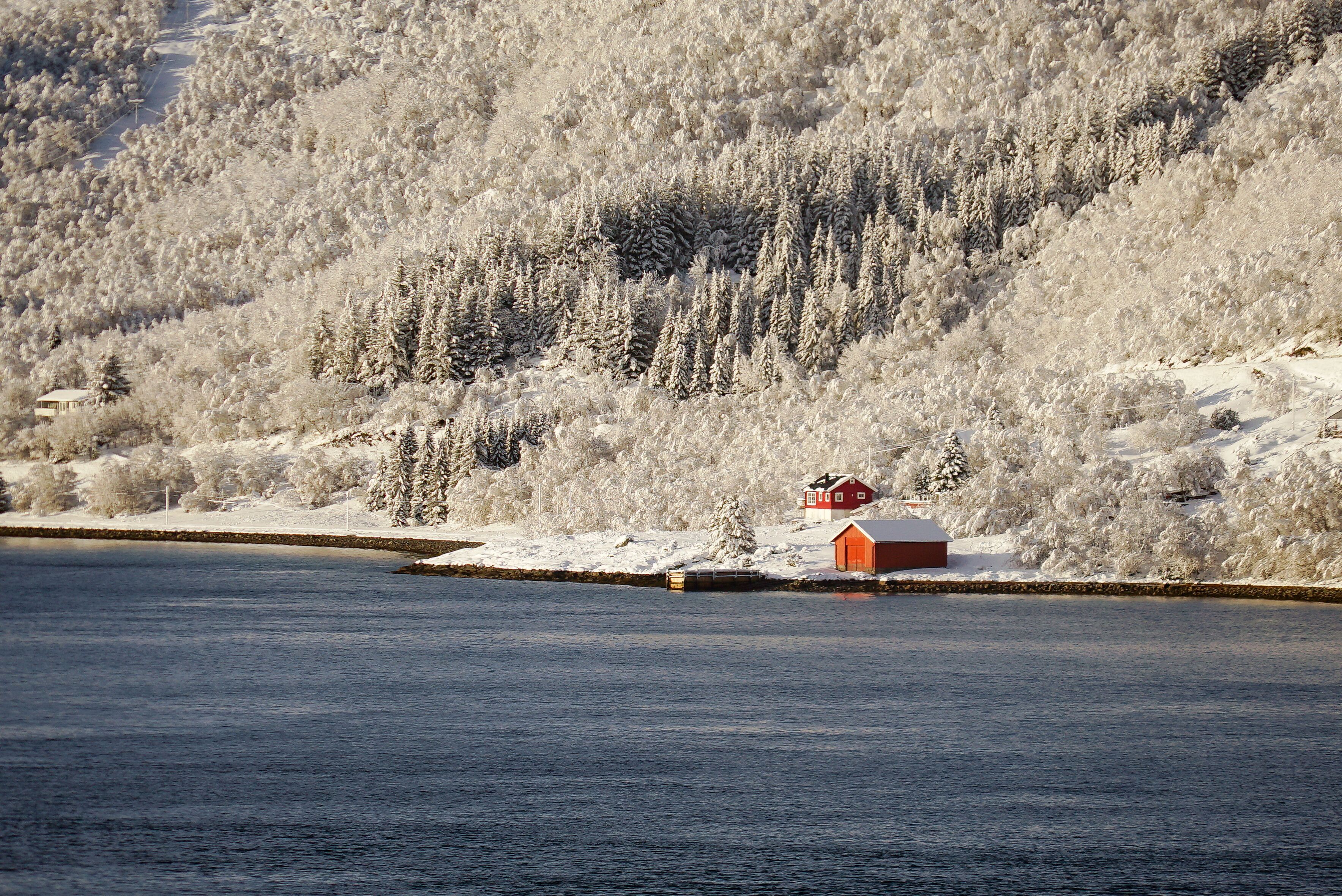 A snow-covered landscape with a red cabin by a calm lake and a forested hill in the background.