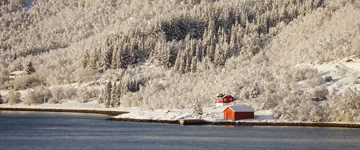 A snow-covered landscape with a red cabin by a calm lake and a forested hill in the background.
