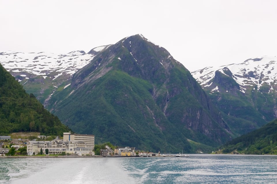 Un fjord avec une montagne en arrière-plan, une petite ville avec des bâtiments le long du rivage et des sommets enneigés.