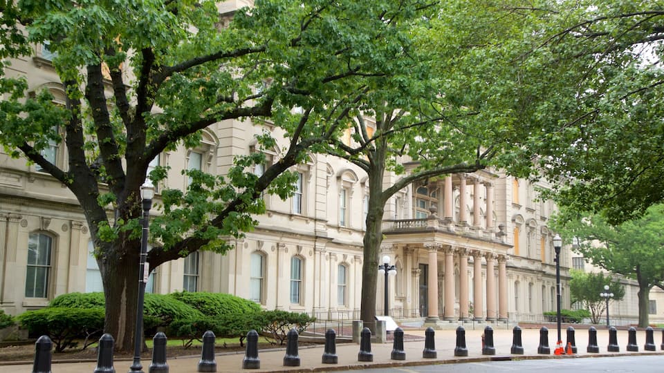 A historic building with classical architecture, surrounded by mature trees and a row of bollards along the sidewalk.