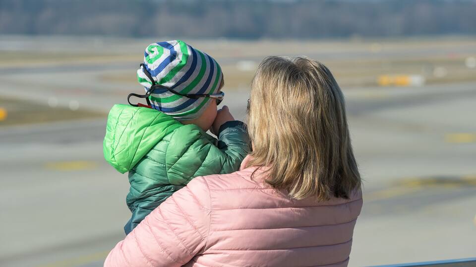 A person in a pink jacket holding a child in a green jacket and striped hat, overlooking an airport runway.