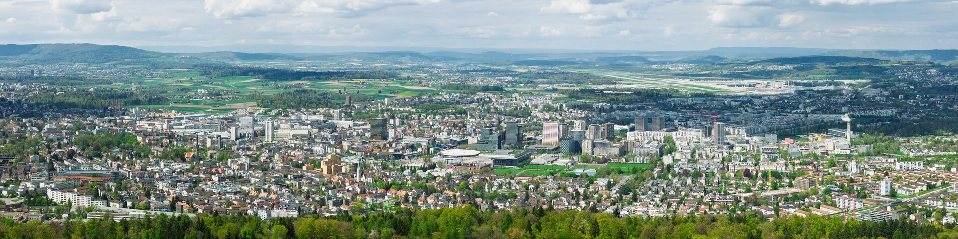 A cityscape with dense urban buildings, a prominent stadium, and a distant airport.