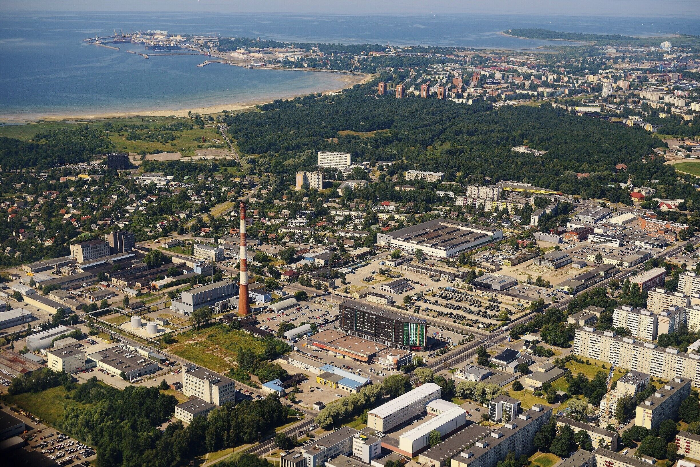 An aerial view of a coastal city with a mix of residential, commercial, and industrial areas, including a prominent factory and a beach in the background.