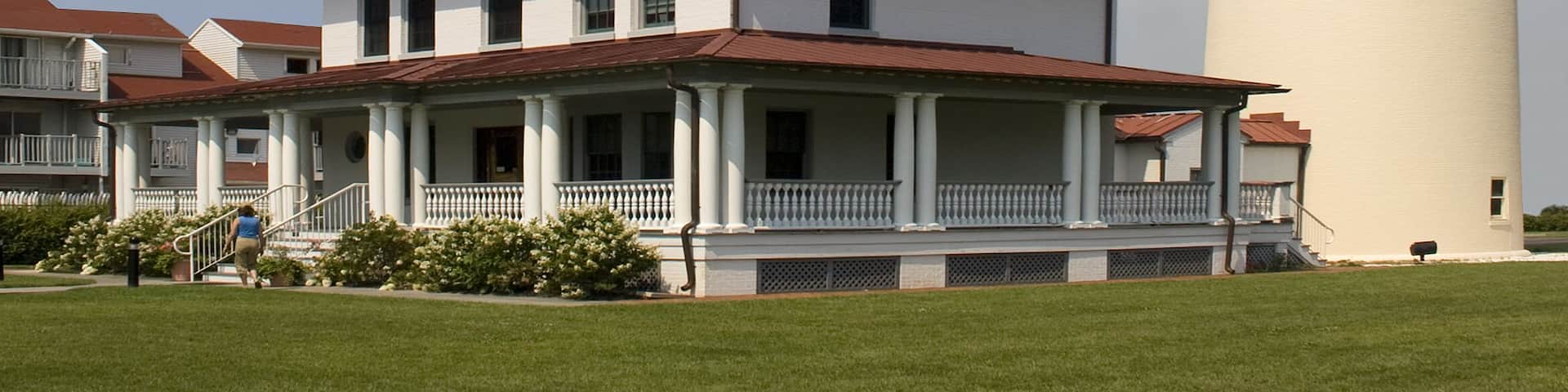 A tall white and black lighthouse with a red roof, a white building with a porch, and a clear blue sky with clouds.