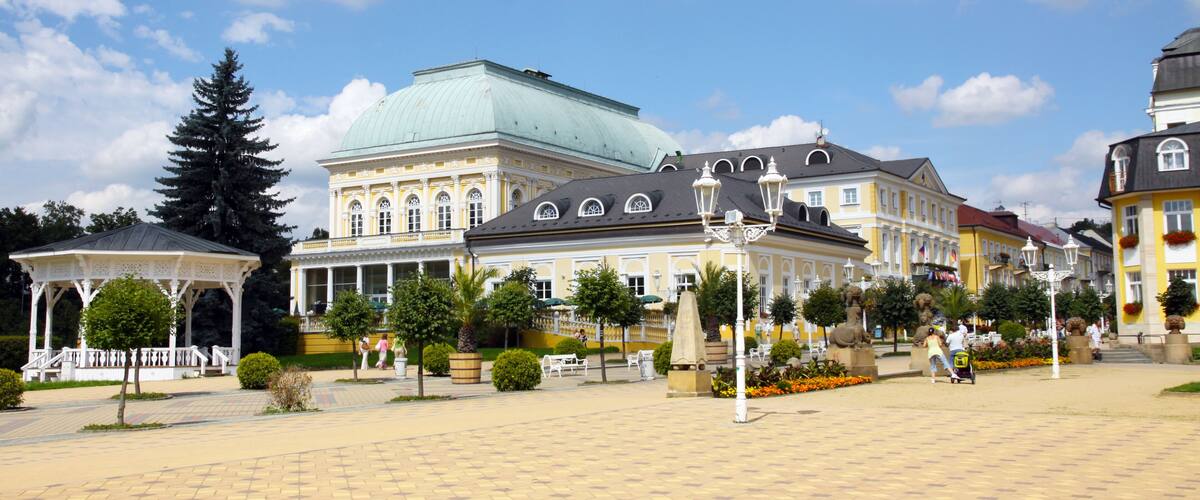 A historic square with a prominent building featuring a green dome, a gazebo, and a statue.