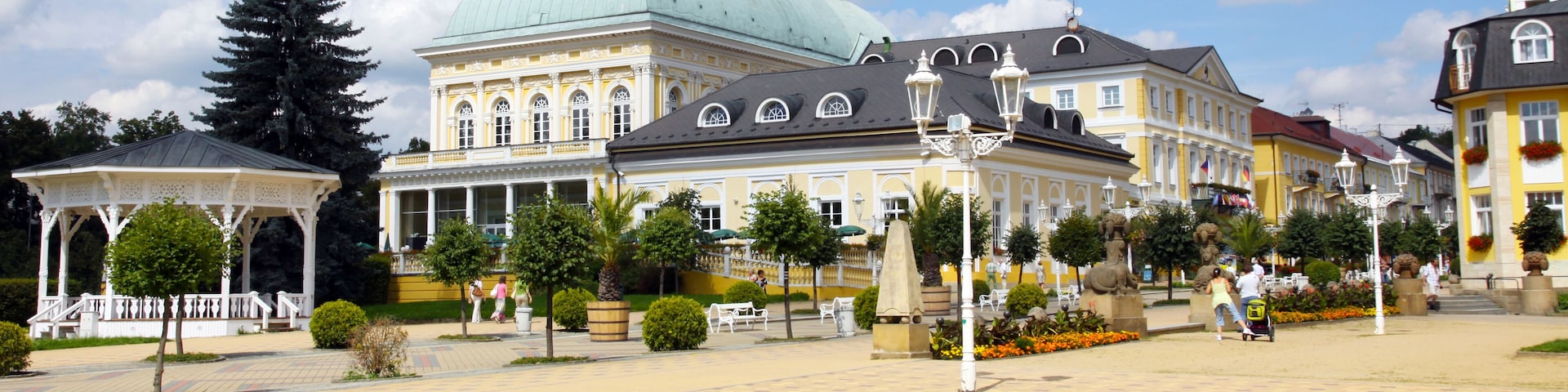 A historic square with a prominent building featuring a green dome, a gazebo, and a statue.