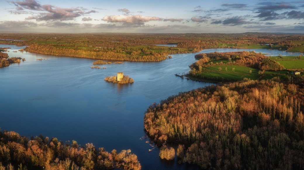 Aerial view of Clough Oughter Castle on a small island in Lough Oughter, surrounded by vibrant autumn forests and calm blue water under a cloudy sky at sunset in County Cavan, Ireland.
