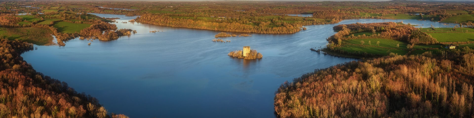 Aerial view of Clough Oughter Castle on a small island in Lough Oughter, surrounded by vibrant autumn forests and calm blue water under a cloudy sky at sunset in County Cavan, Ireland.