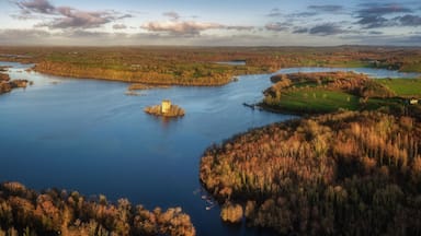 Aerial view of Clough Oughter Castle on a small island in Lough Oughter, surrounded by vibrant autumn forests and calm blue water under a cloudy sky at sunset in County Cavan, Ireland.