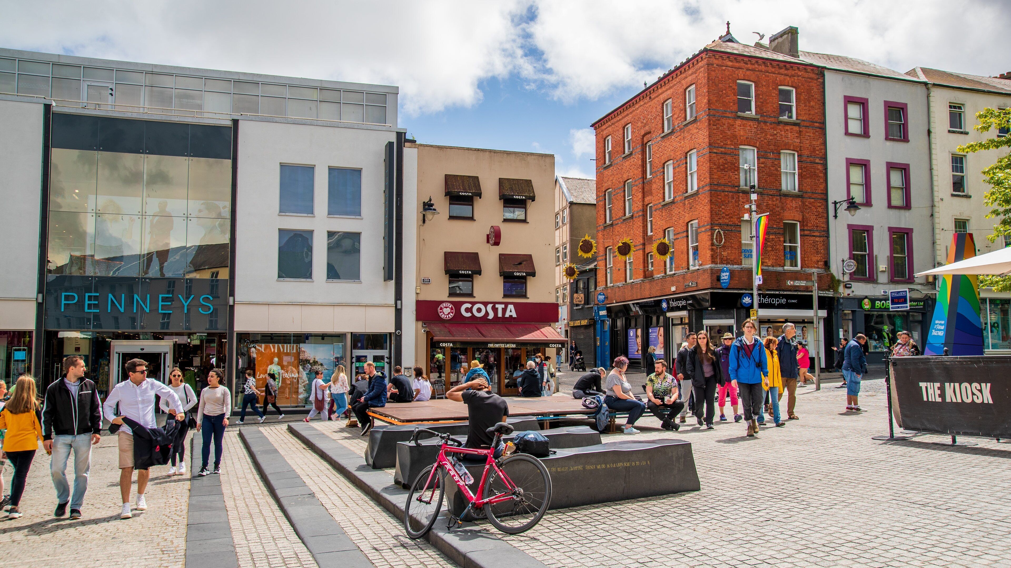 Una calle urbana concurrida con tiendas como Costa y Penneys, una bicicleta roja y personas caminando.