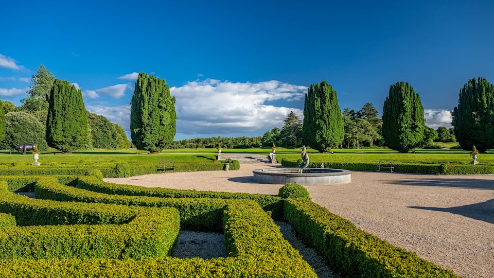 A formal garden with neatly trimmed hedges, tall cypress trees, and a central circular fountain.