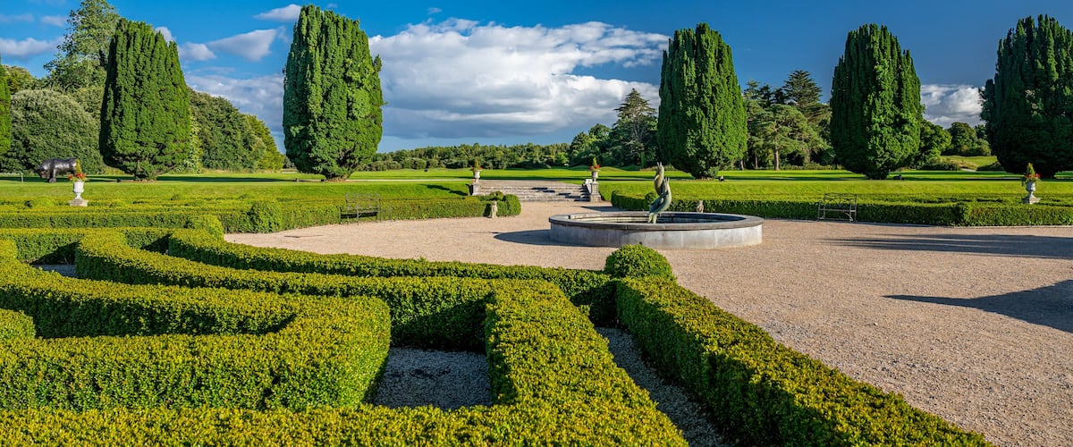 A formal garden with neatly trimmed hedges, tall cypress trees, and a central circular fountain.